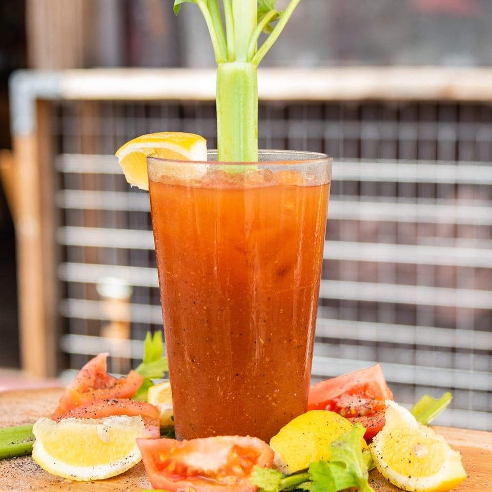 Bloody Mary cocktail garnished with celery and lemon, surrounded by sliced tomatoes, citrus, and fresh vegetables on a wooden table.