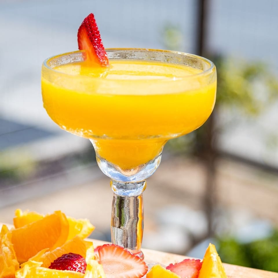 Bright orange cocktail in a margarita glass garnished with a strawberry, surrounded by sliced oranges and strawberries.