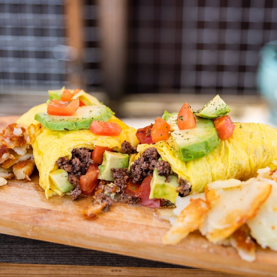 Breakfast omelets filled with ground beef, avocado, and tomatoes served alongside crispy potatoes on a wooden board.